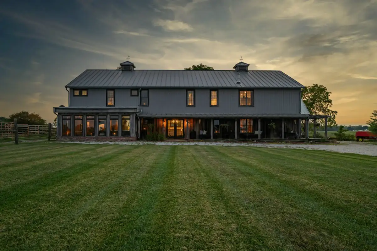 A long two-story farmhouse with a standing-seam metal roof and wraparound porch, lit at dusk across a wide lawn