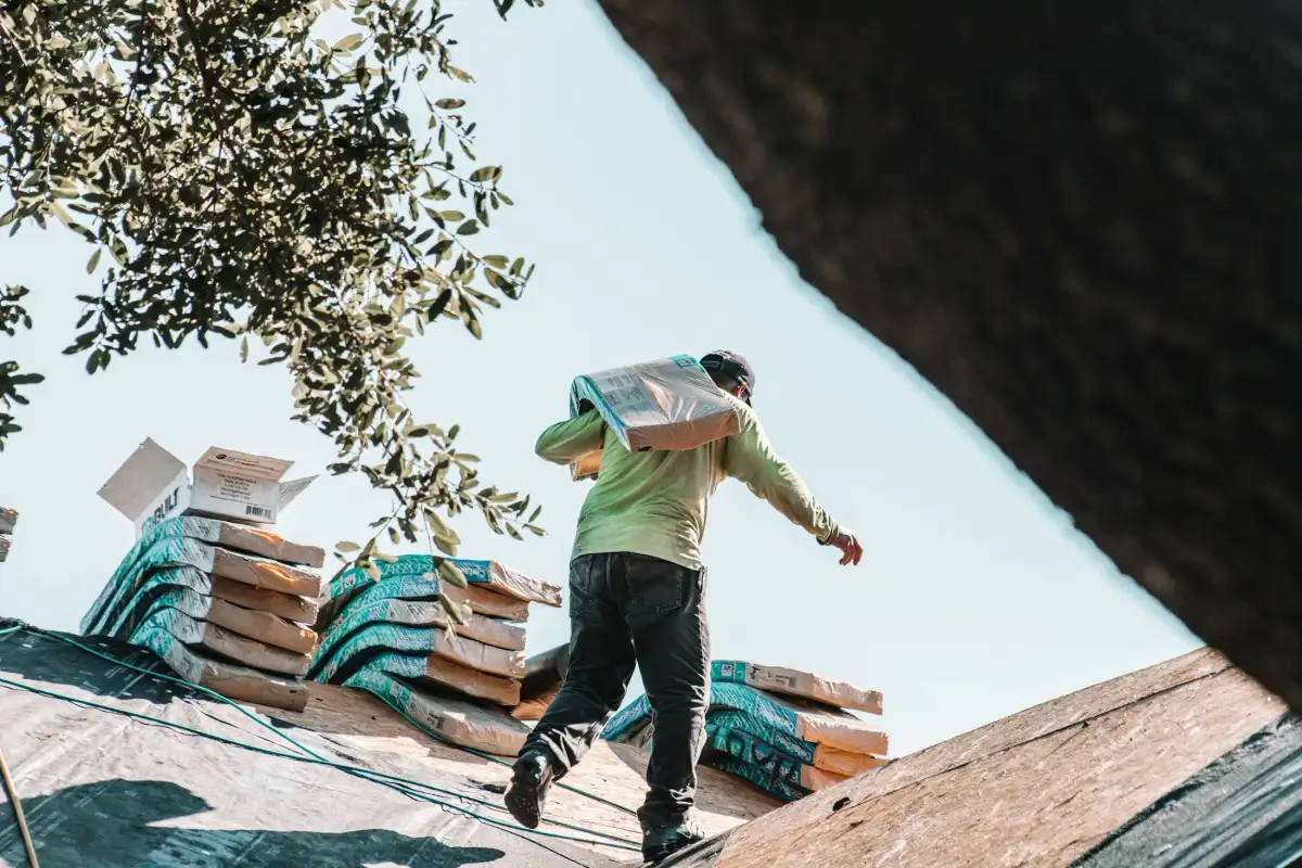 A worker carries a bundle of shingles across a roof deck past stacked bundles