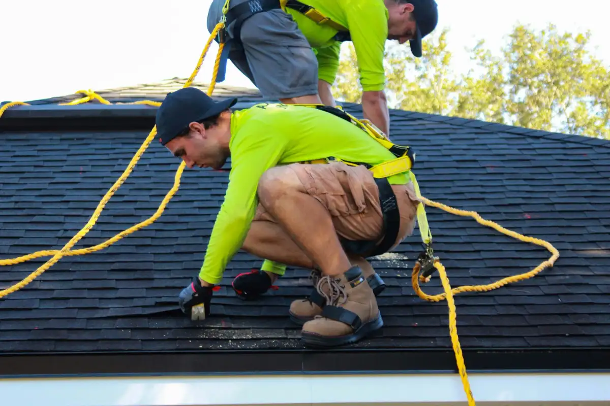 Two roofers in hi-vis shirts and harnesses tied off on a dark shingle roof