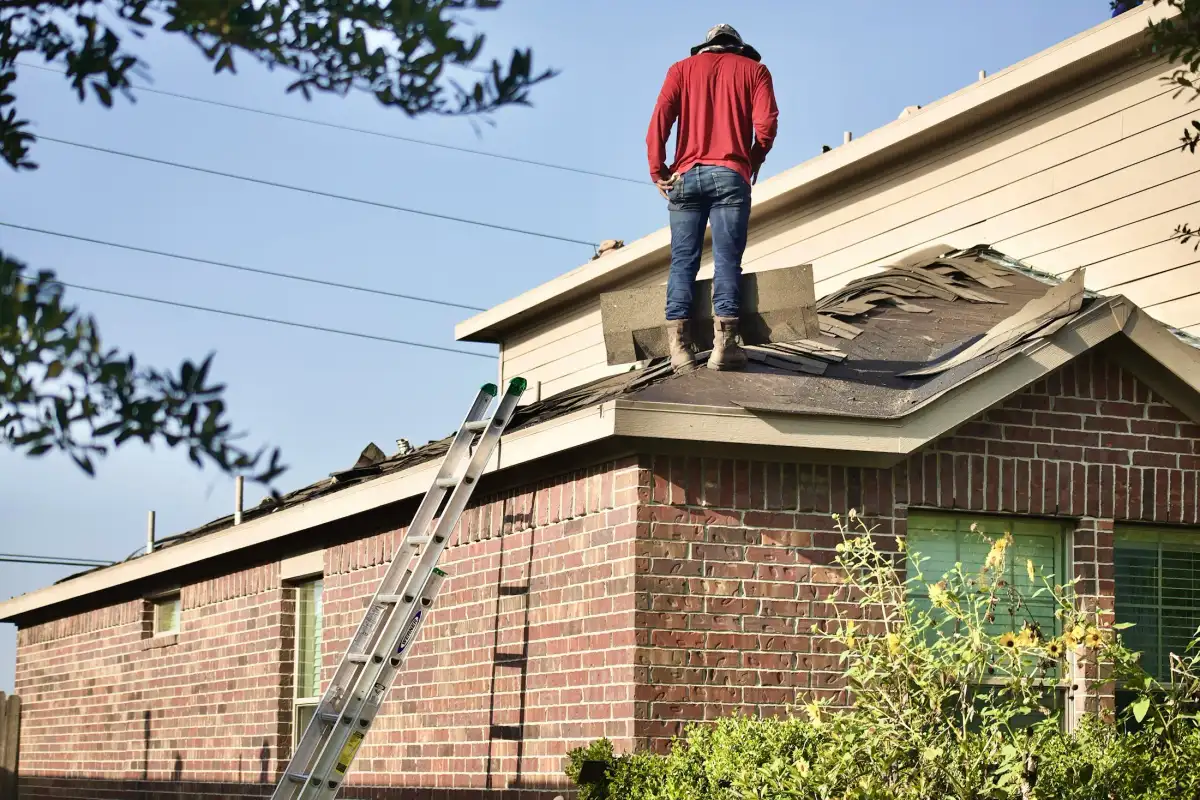 A roofer in a red shirt stands on a brick house mid tear-off, old shingles piled on the bare deck, a ladder against the wall