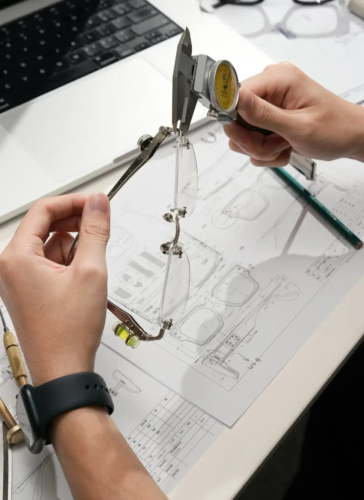 Hands measuring a rimless frame with a dial caliper over technical drawings (stock photo)